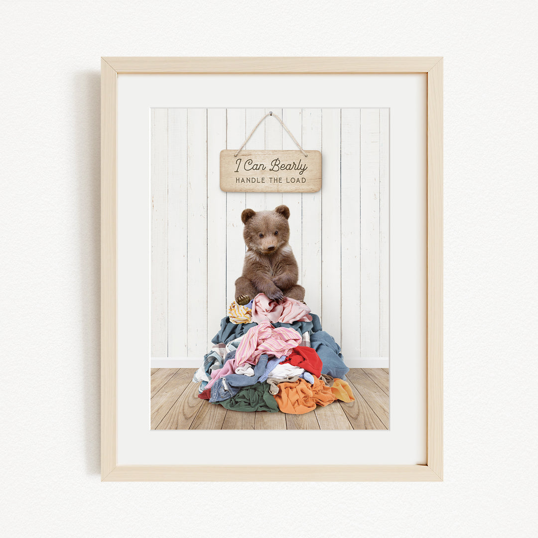 A framed image of a brown bear sitting atop a pile of clothes, with a wooden sign above that reads "I Can Bearly Handle the Load".