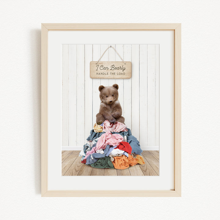 A framed image of a brown bear sitting atop a pile of clothes, with a wooden sign above that reads "I Can Bearly Handle the Load".