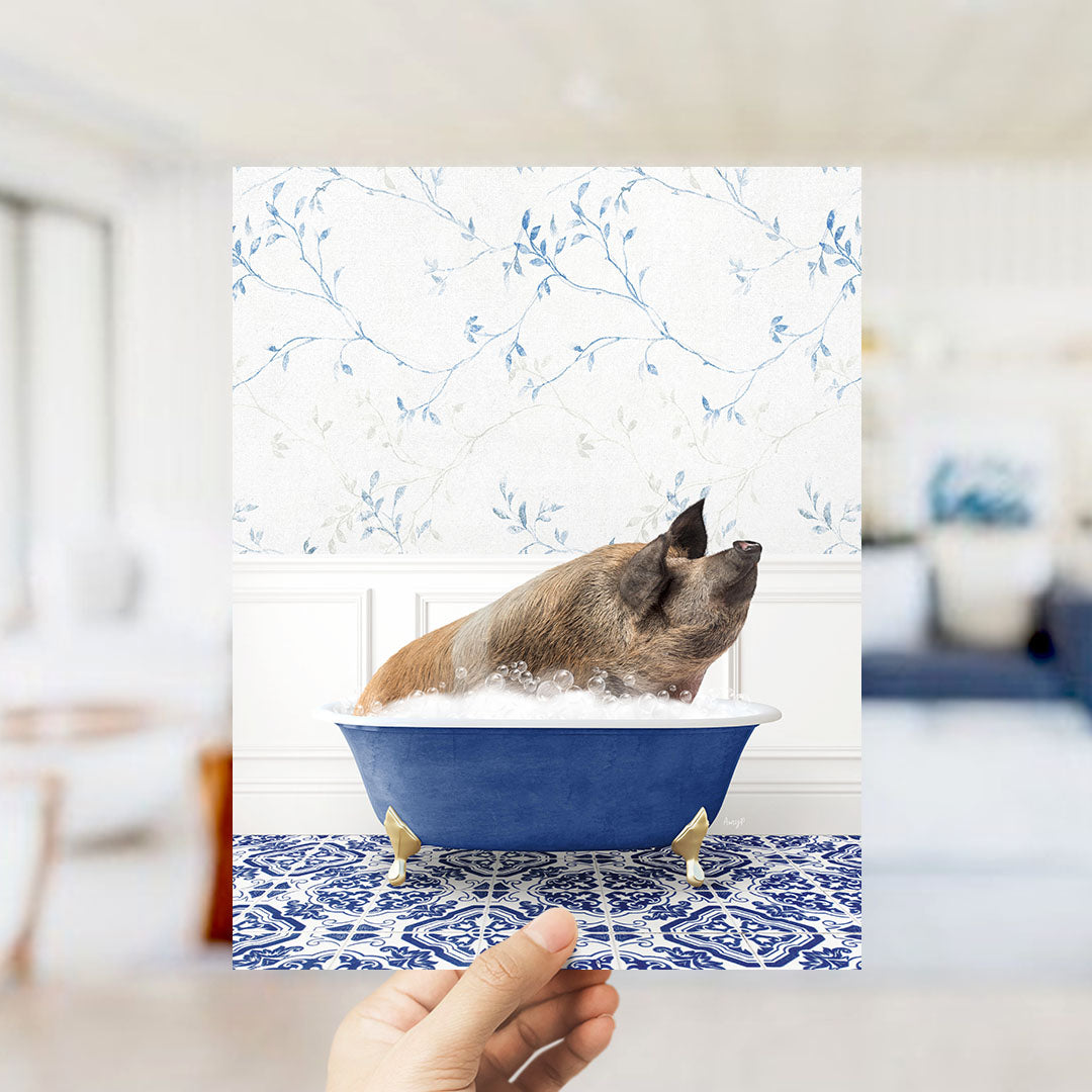 A hand holds up a framed image of a pig sitting in a blue bathtub filled with bubbles, against a backdrop of a blue and white floral wallpaper.