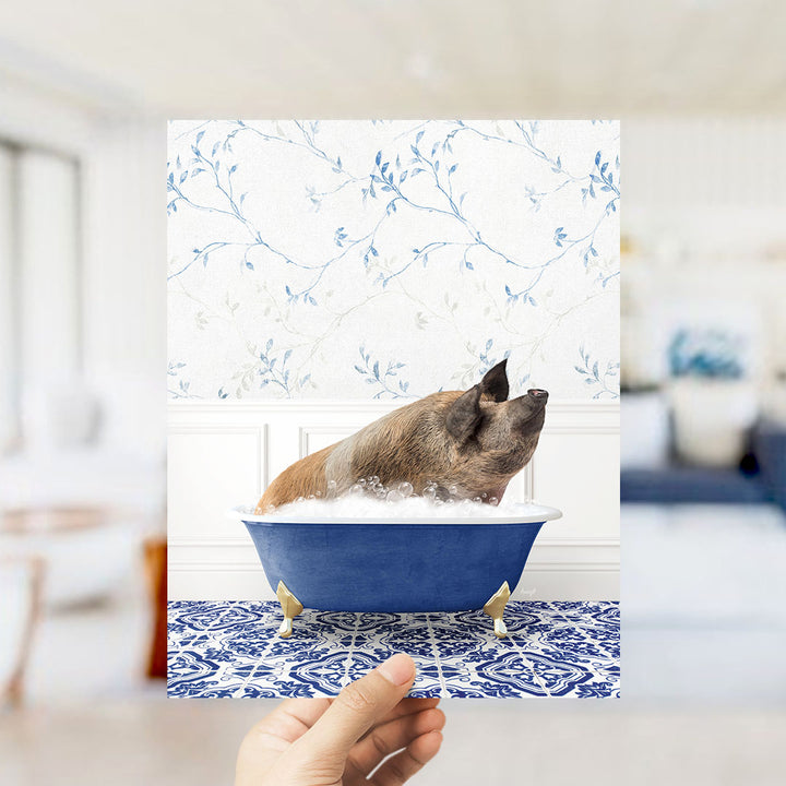 A hand holds up a framed image of a pig sitting in a blue bathtub filled with bubbles, against a backdrop of a blue and white floral wallpaper.