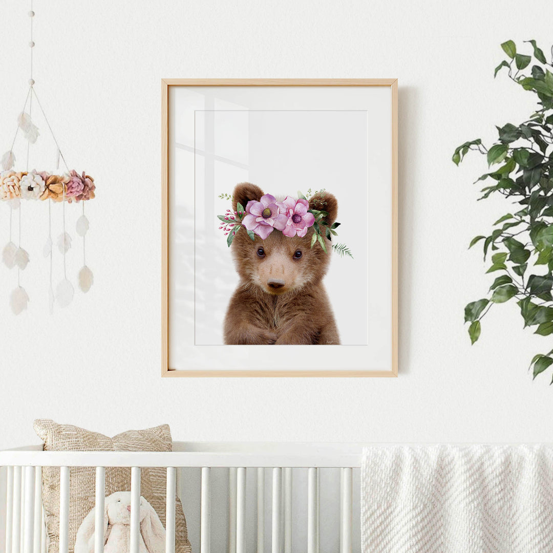 A framed image of a brown bear wearing a floral crown is displayed on a white wall, with a crib and a hanging mobile with flowers in the background.