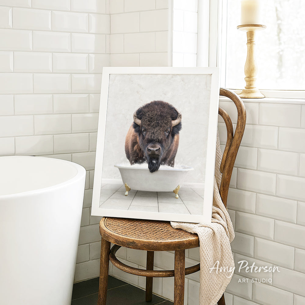 A framed image of a bison sitting in a bathtub is displayed on a wooden chair in a bathroom setting.
