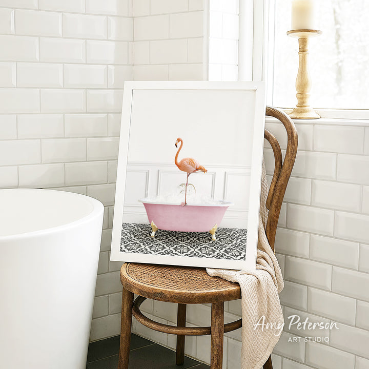 A framed image of a pink flamingo sitting in a pink bathtub is displayed on a wooden chair in a bathroom setting.