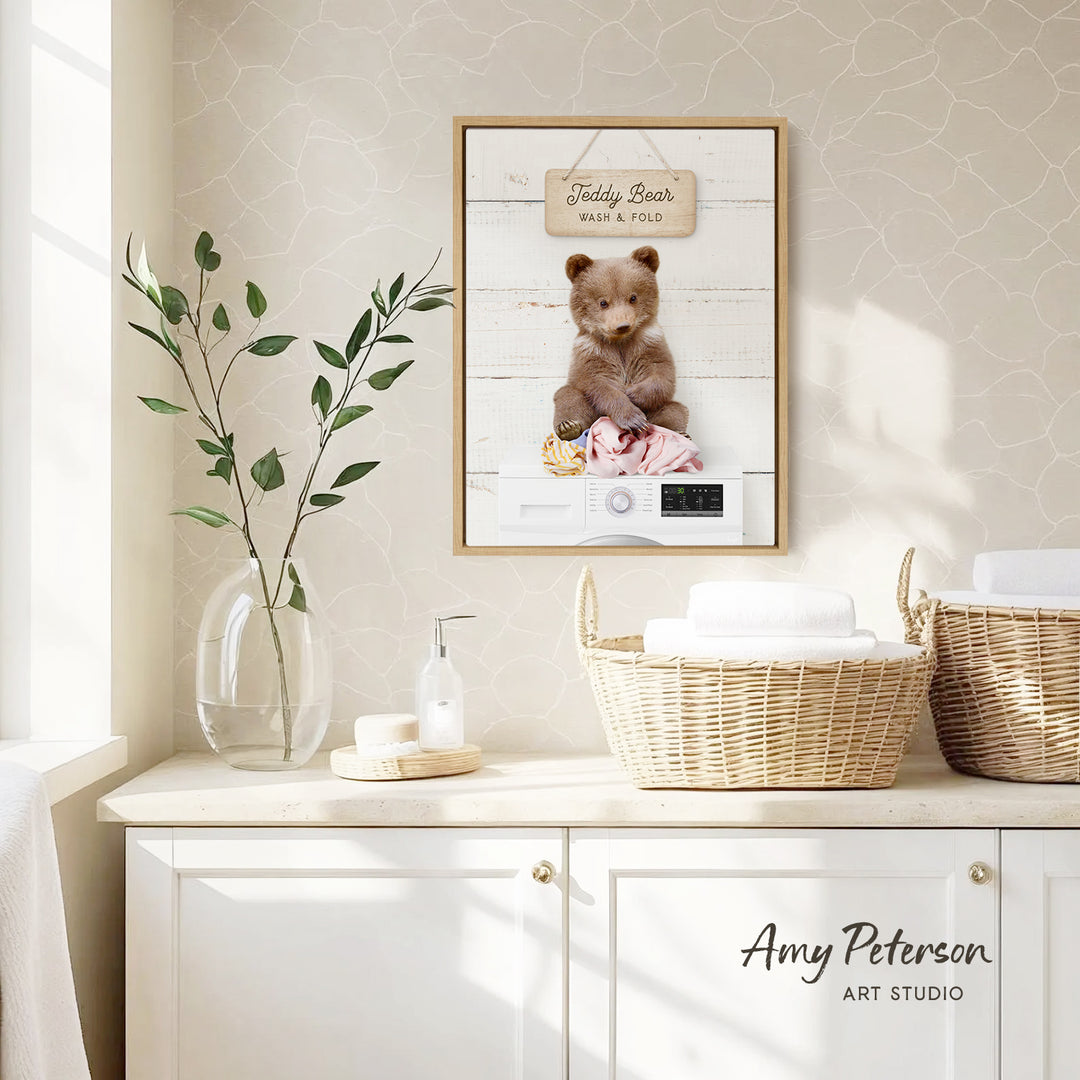 a framed photo of a teddy bear sitting in a basket, with the text "Teddy Bear Wash & Fold" on the frame. The frame is hanging on a wall above a white cabinet, which has a vase of leaves and a bottle of soap on it.