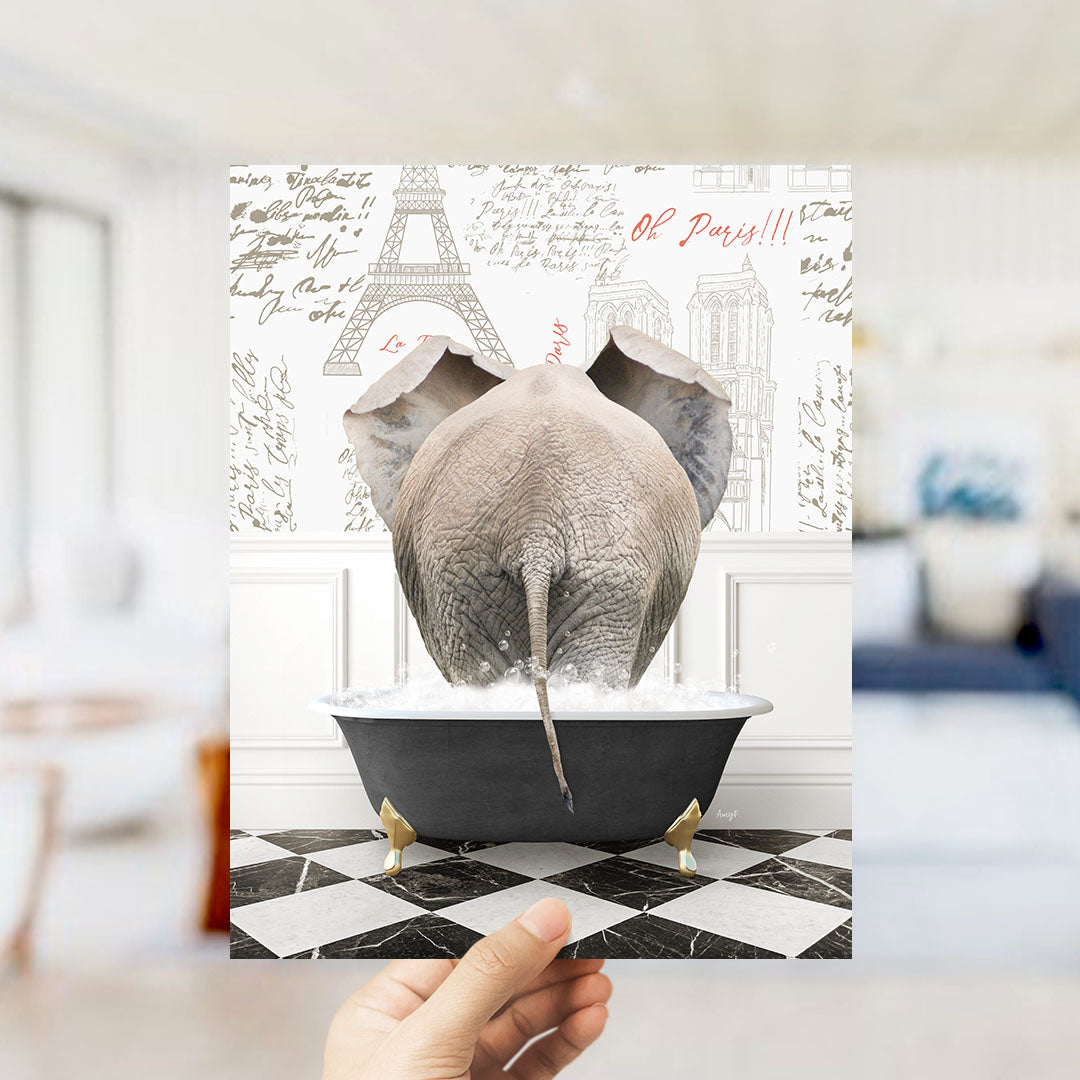 A hand is holding up a framed image depicting an elephant sitting in a bathtub filled with water, with the Eiffel Tower and Paris written on the wall behind it.