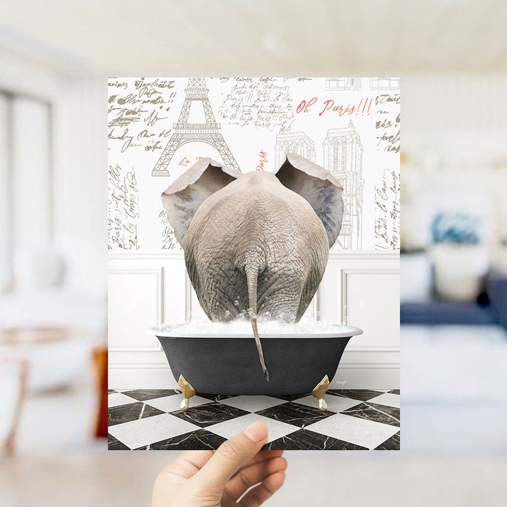 A hand is holding up a framed image depicting an elephant sitting in a bathtub filled with water, with the Eiffel Tower and Paris written on the wall behind it.