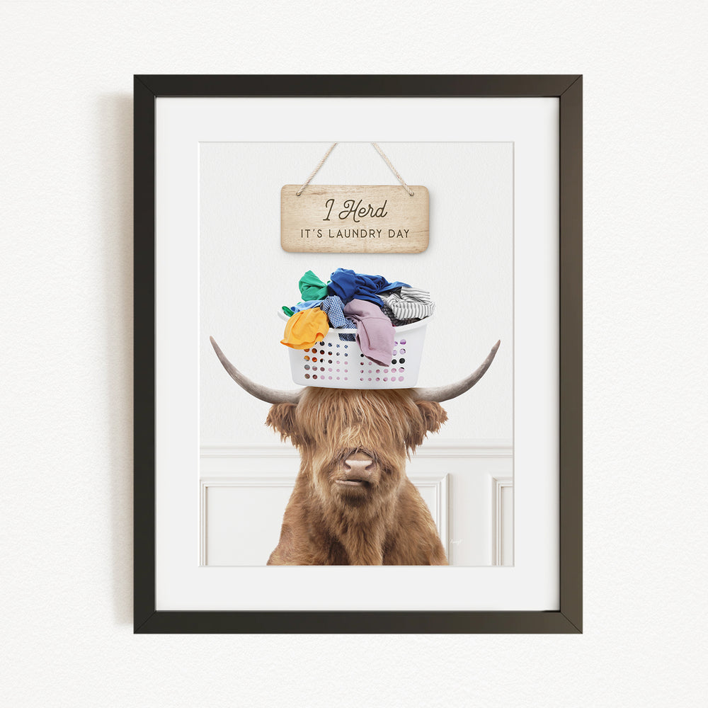 A framed image of a Highland cow with a basket of laundry on its head, accompanied by a wooden sign that reads "I herd it's laundry day".