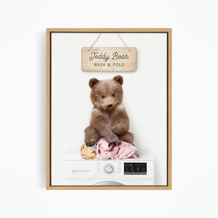 A teddy bear sitting on a pile of laundry inside a washing machine, with a wooden sign above that says "Teddy Bear WASH & FOLD".