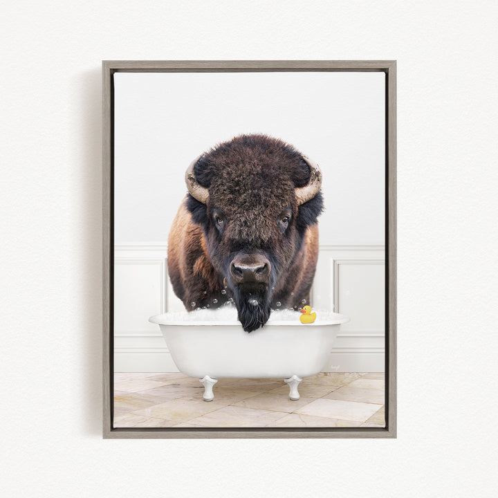 A buffalo with large horns is standing in a white bathtub filled with bubbles, with a rubber duck floating in the water.