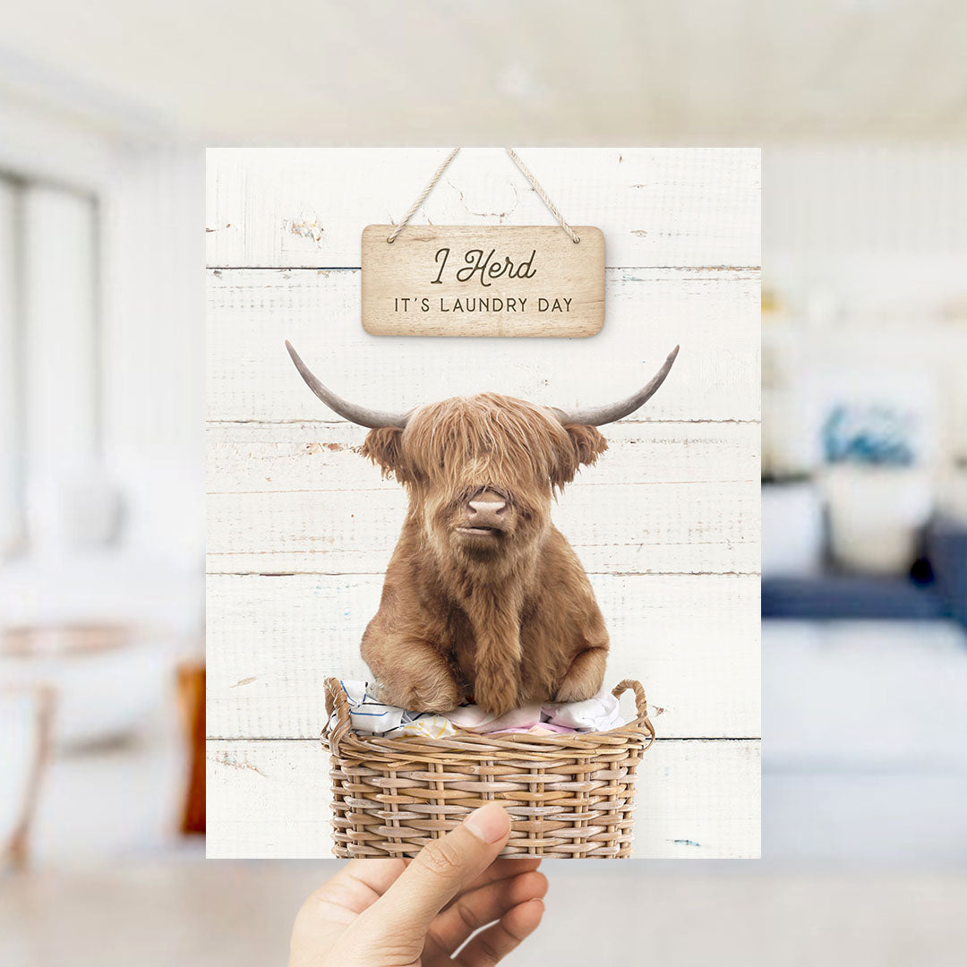 A hand is holding up a card with a picture of a brown cow with long horns sitting in a wicker basket, with a sign above it that says "I herd it's laundry day".