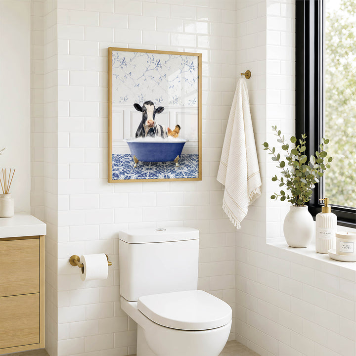 A bathroom with a white toilet, a framed picture of a cow in a bathtub, and a window with greenery outside.