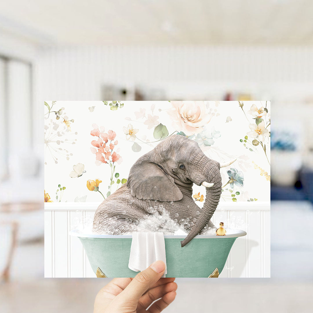 A hand is holding up a framed image of an elephant sitting in a bathtub filled with water and surrounded by flowers.