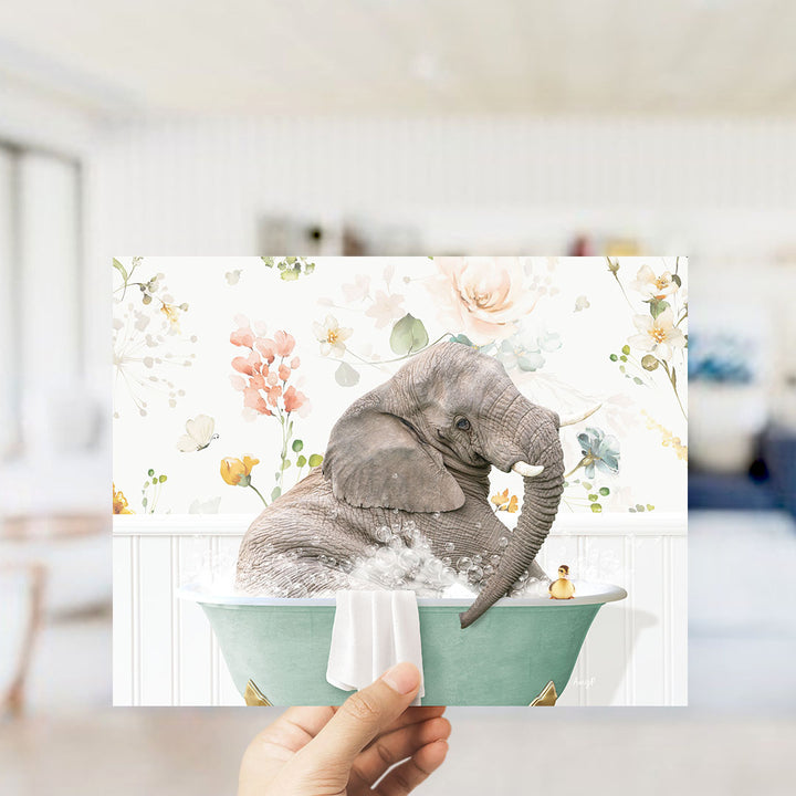 A hand is holding up a framed image of an elephant sitting in a bathtub filled with water and surrounded by flowers.