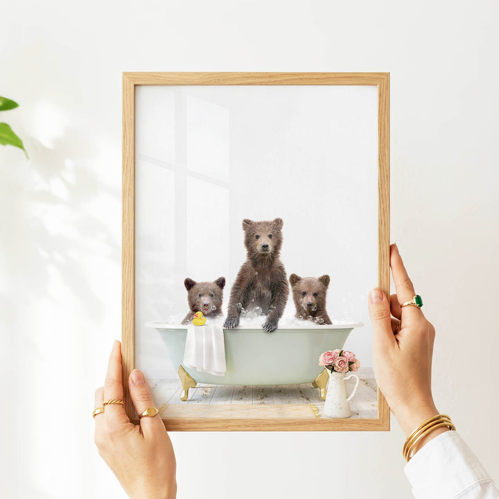 A person's hands are holding a picture frame with a framed image of three bears sitting in a bathtub.