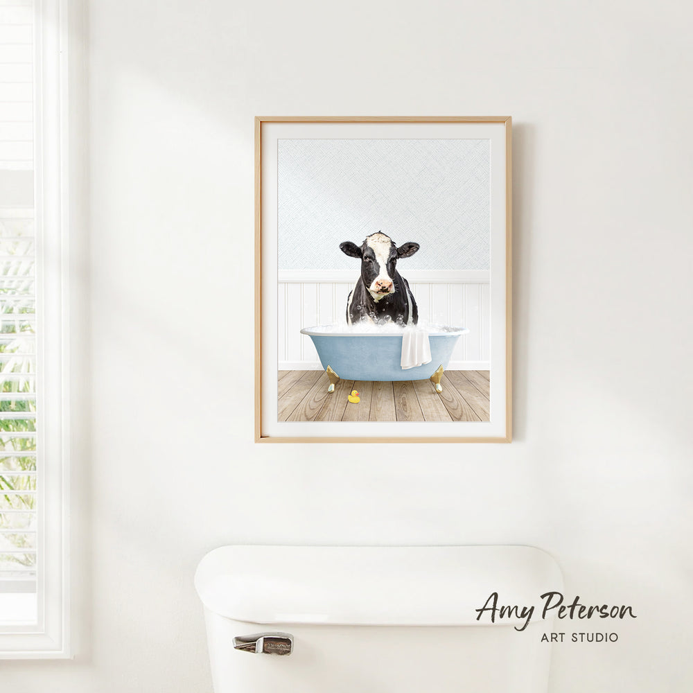 A framed print of a cow sitting in a bathtub is hanging on a white wall.