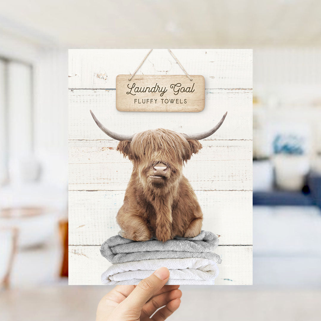 A hand is holding up a sign with a picture of a brown cow with large horns, sitting on top of a stack of towels.