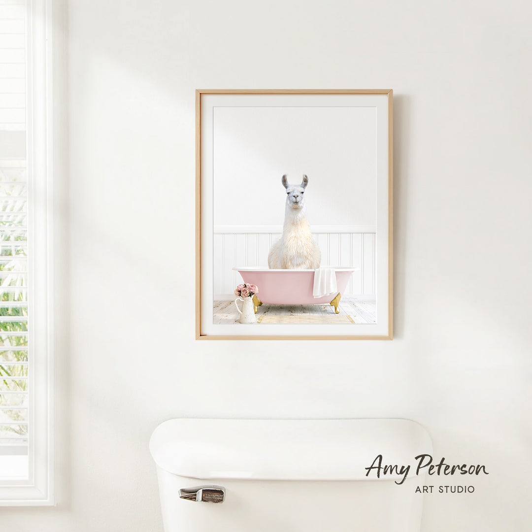 A framed image of a white llama sitting in a pink bathtub is hanging on a white wall in a bathroom.