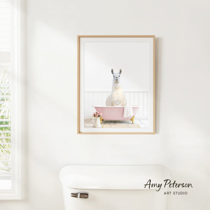 A framed image of a white llama sitting in a pink bathtub is hanging on a white wall in a bathroom.