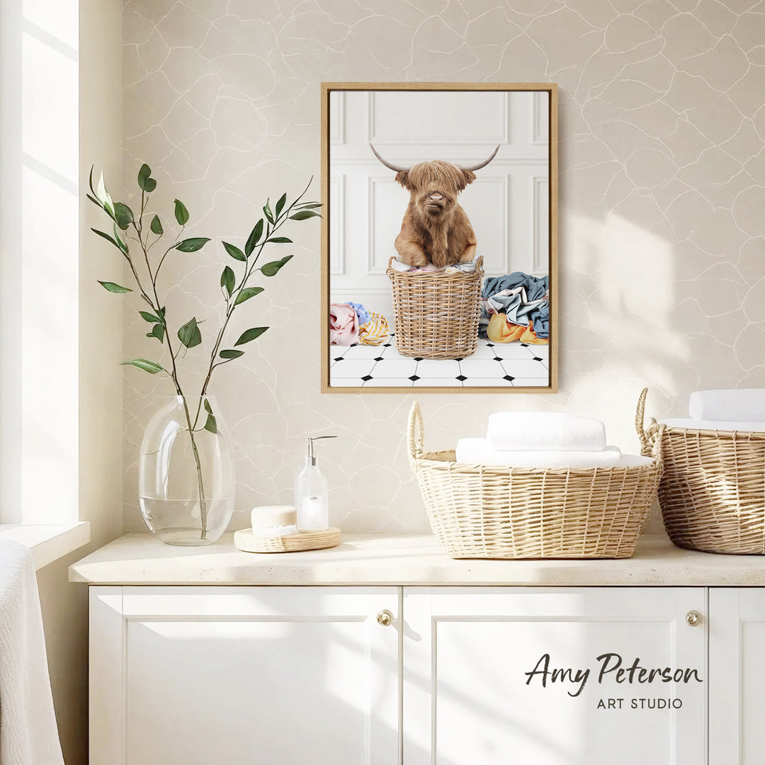 a framed photograph of a Highland cow sitting in a wicker basket, placed on a wall above a white cabinet with two woven baskets on top.