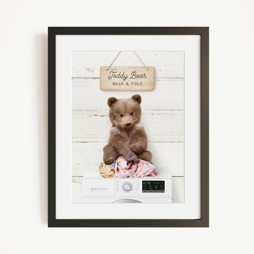 A framed image of a teddy bear sitting on a washing machine, with a sign above it that says "Teddy Bear Wash & Fold".
