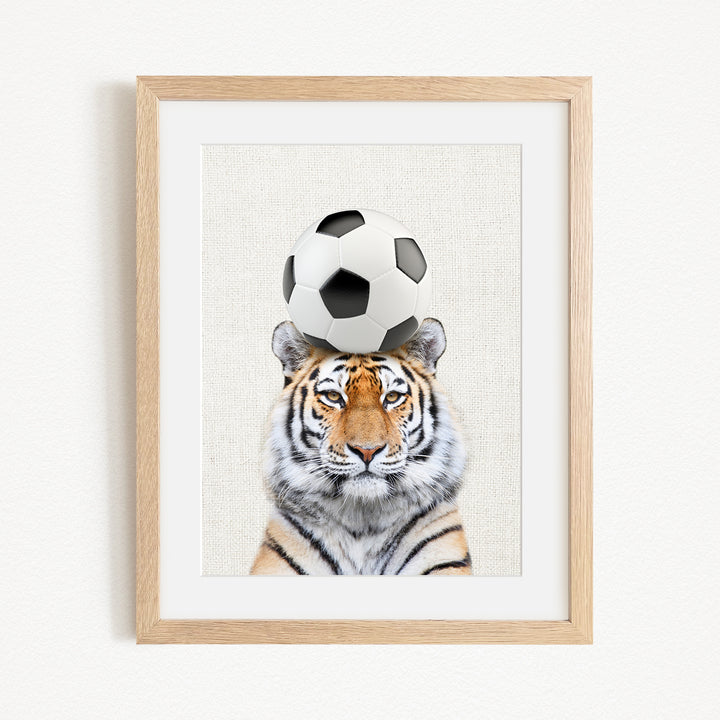 A tiger is sitting in front of a soccer ball, with the ball resting on its head.