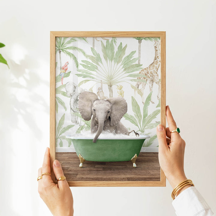 A person's hands are holding a framed picture of an elephant sitting in a green bathtub, surrounded by tropical foliage.