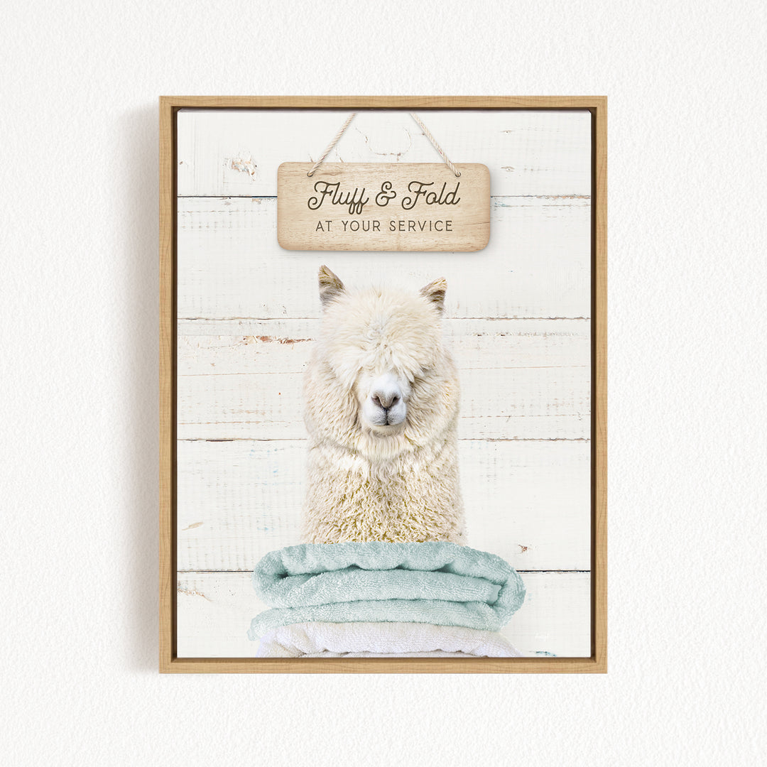 A fluffy white alpaca sitting on a stack of light blue towels, with a wooden sign above that reads "Fuffy & Fold at your Service".