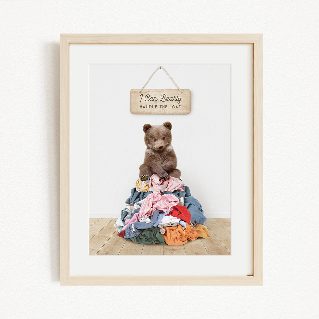A framed image of a brown bear sitting atop a pile of clothes, with a wooden sign above that reads "I Can Bearly Handle the Load".