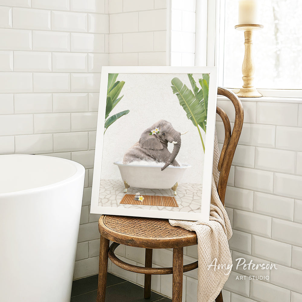 A framed image of a gray elephant sitting in a white bathtub, surrounded by green plants, is displayed on a wooden chair in a bathroom setting.