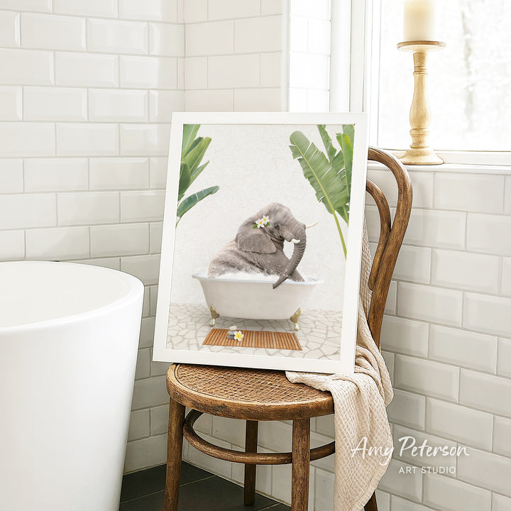 A framed image of a gray elephant sitting in a white bathtub, surrounded by green plants, is displayed on a wooden chair in a bathroom setting.