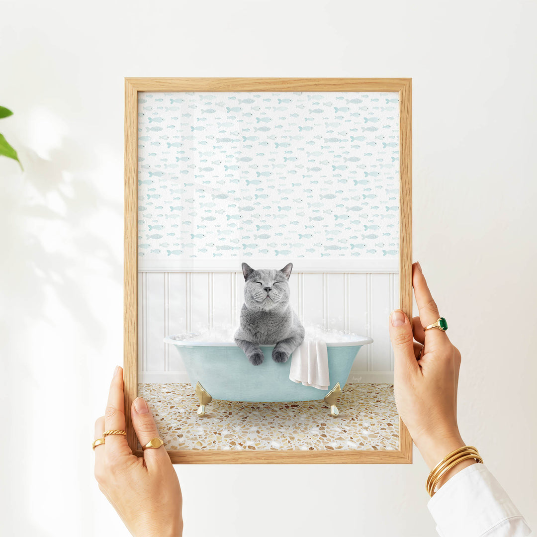 A person's hands are holding a framed picture of a gray cat sitting in a blue bathtub filled with bubbles, with a fish-themed wallpaper in the background.