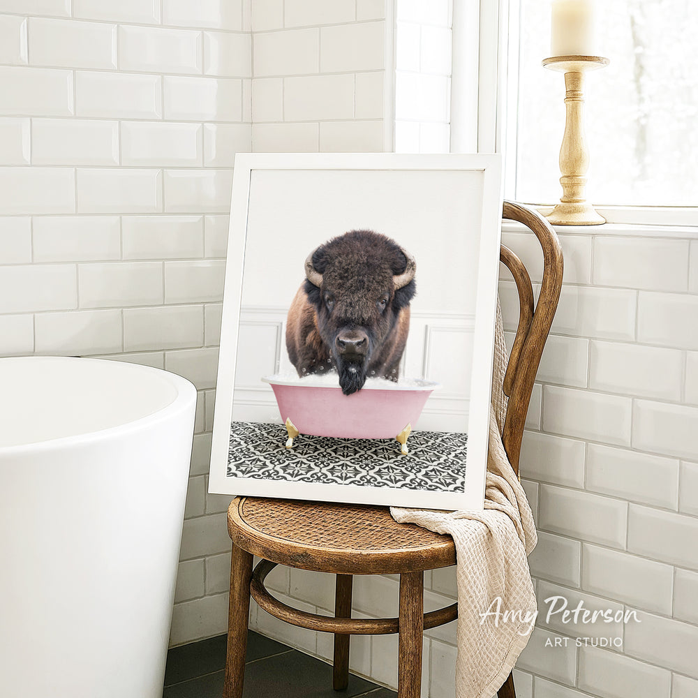 A framed image of a bison sitting in a pink bathtub is displayed on a wooden chair in a bathroom.