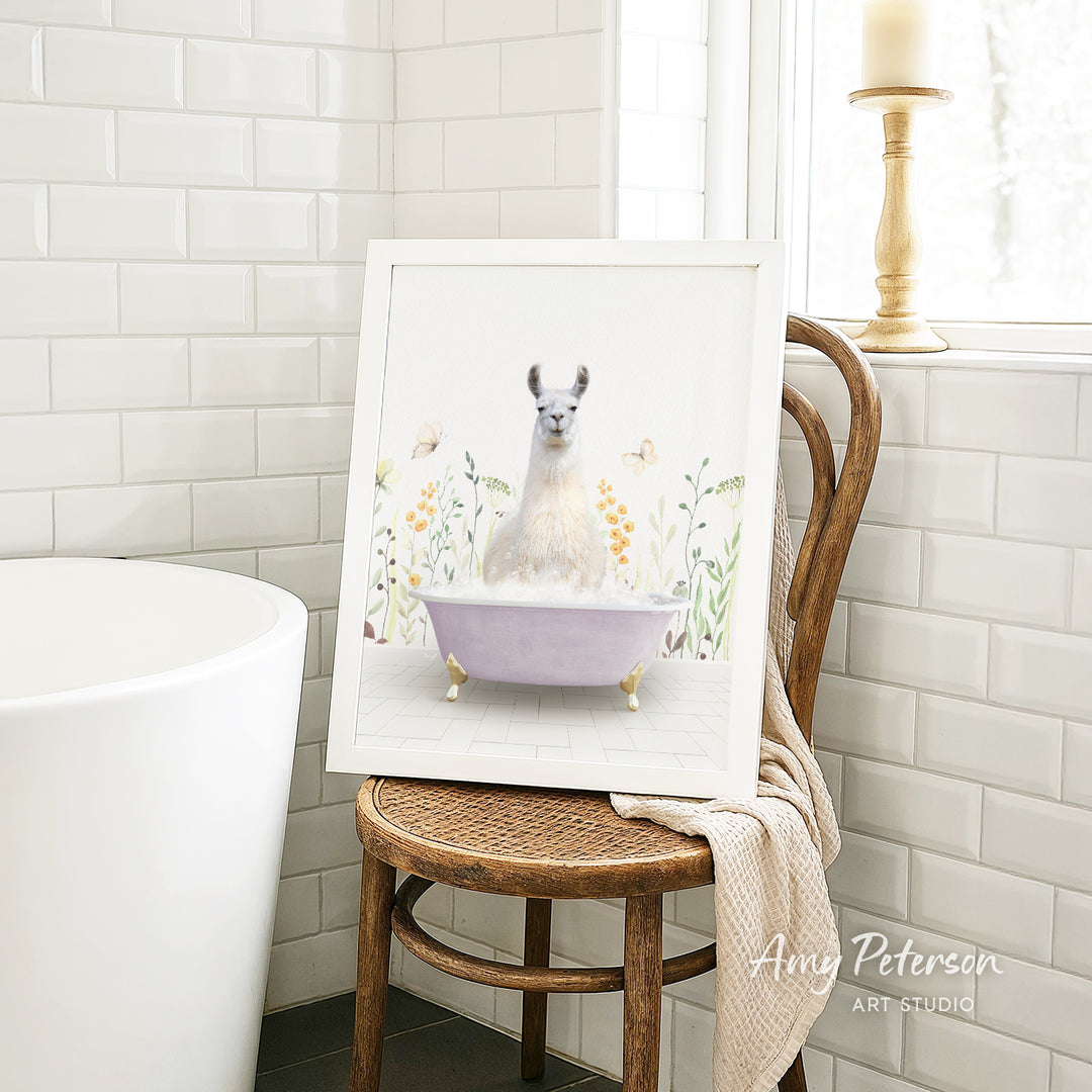 A framed image of a white llama sitting in a purple bathtub, surrounded by flowers, is displayed on a wooden chair in a bathroom setting.
