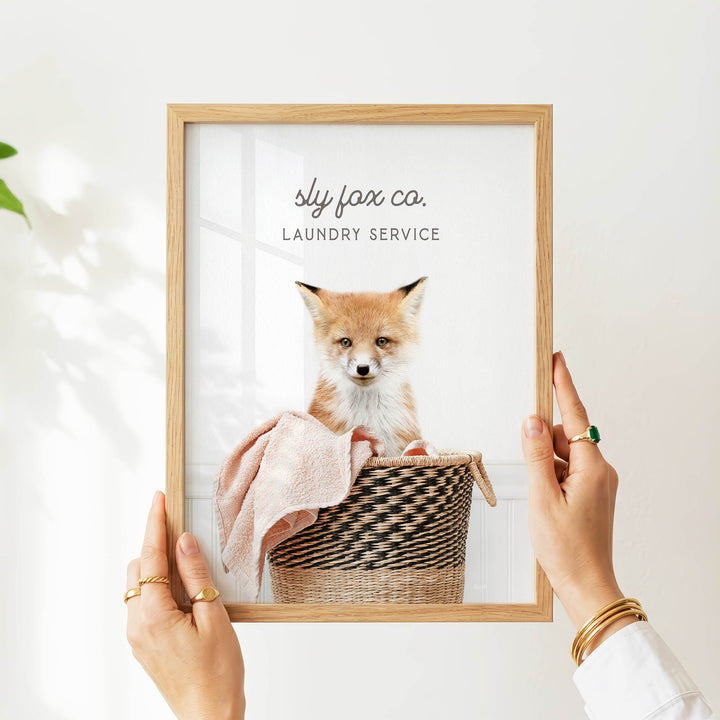 A person is holding a framed picture of a small orange and white fox sitting in a woven basket.