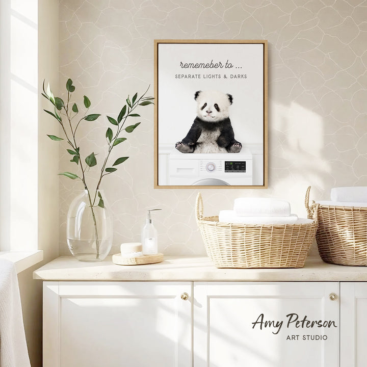 a framed poster of a panda bear sitting on a washing machine, with a wicker basket and a plant on a white countertop below it.