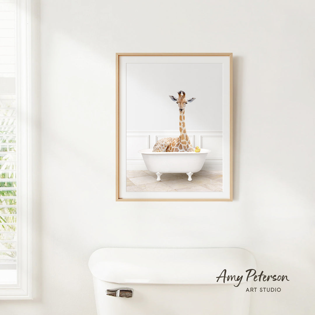 A framed image of a giraffe sitting in a white bathtub, surrounded by various objects, is displayed on a white wall above a white toilet.