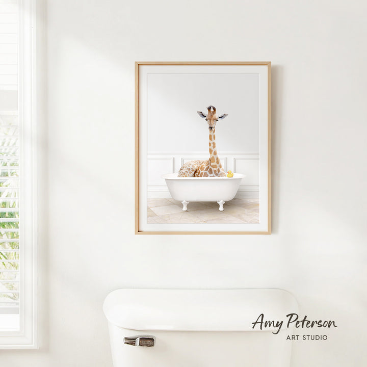 A framed image of a giraffe sitting in a white bathtub, surrounded by various objects, is displayed on a white wall above a white toilet.