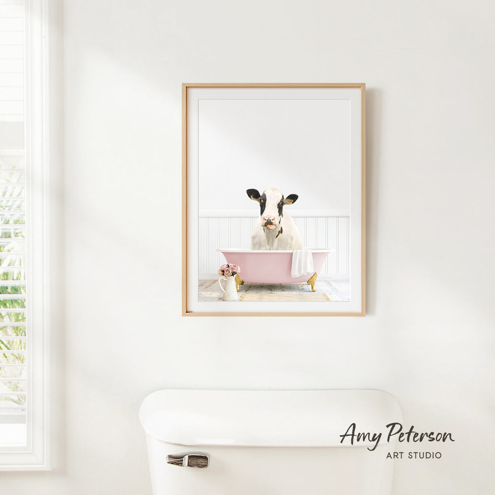 A framed image of a cow sitting in a pink bathtub is hanging on a white wall in a bathroom.