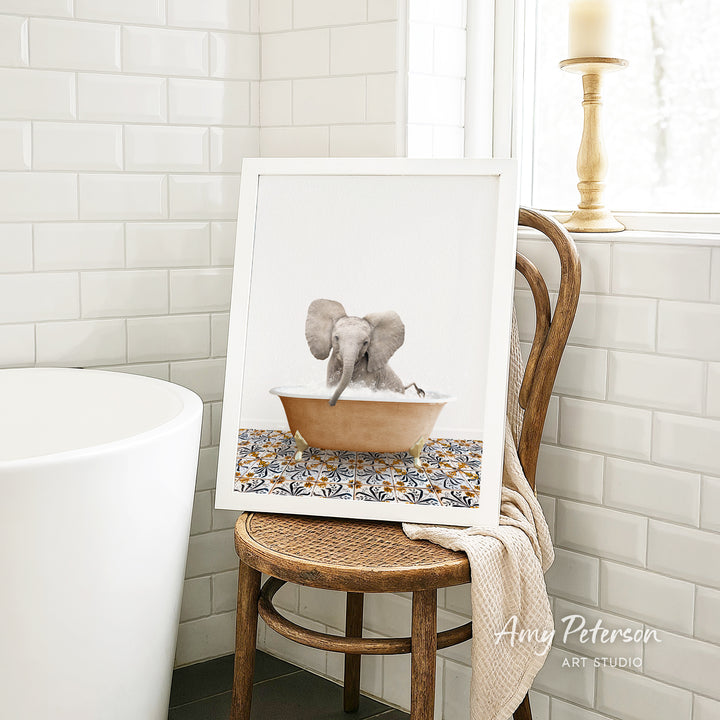A framed image of an elephant sitting in a bathtub, with a towel draped over the back of a wooden chair in a bathroom setting.