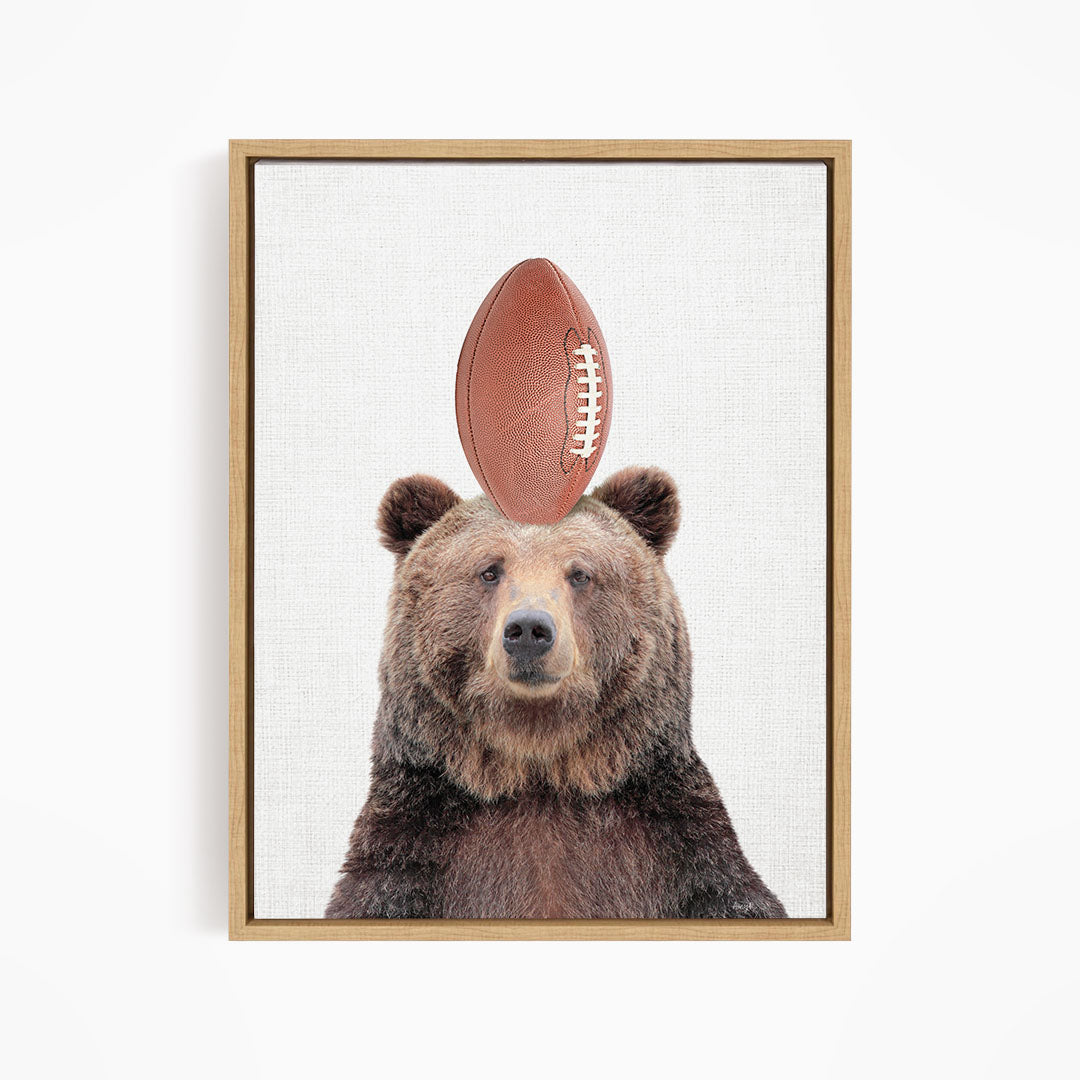 A brown bear with a football on its head, looking directly at the camera.