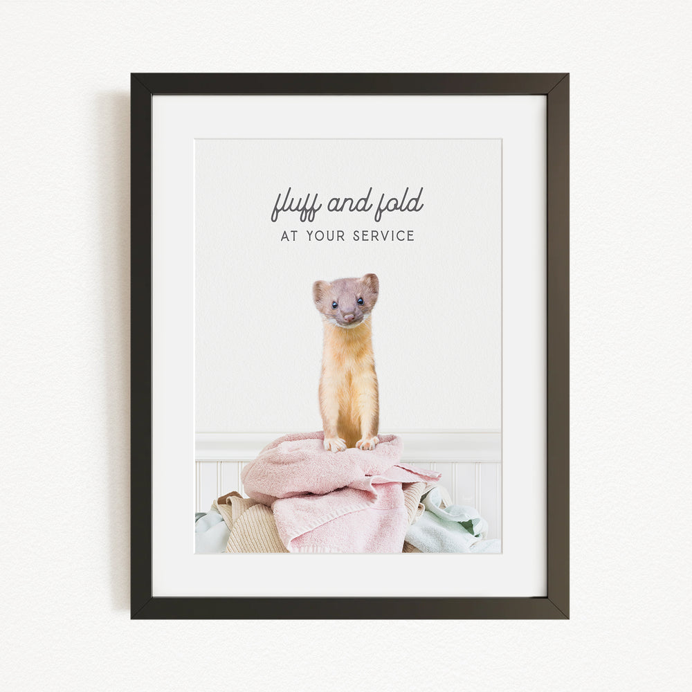A small brown and white ferret sitting on a pile of clothes in a framed picture.