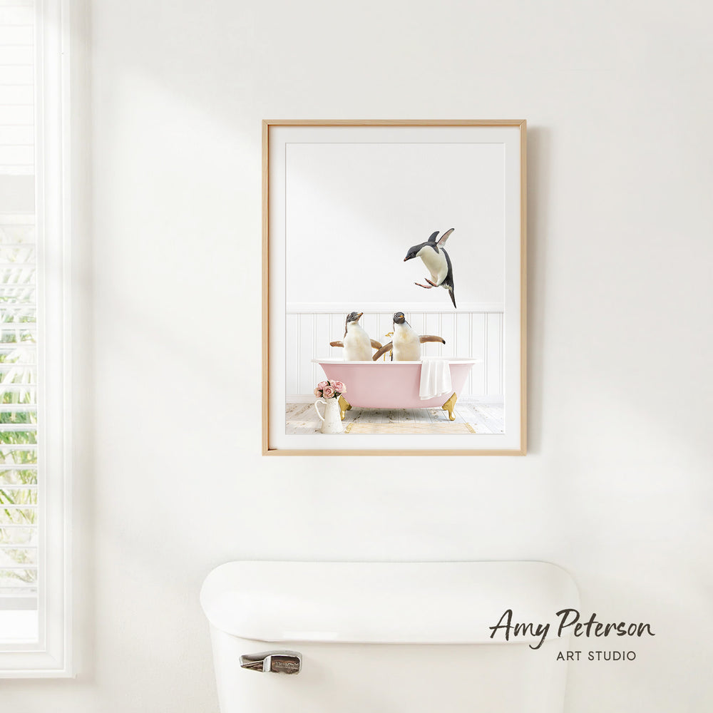 A framed image of two penguins sitting in a pink bathtub, with one penguin flying towards the other, is hanging on a white wall in a bathroom.