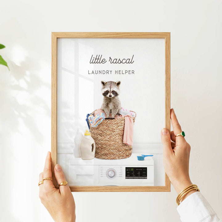 A person is holding up a framed image of a raccoon sitting in a laundry basket, with various household items visible in the basket.