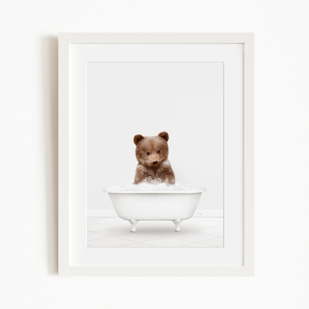 A cute brown bear sitting in a white bathtub filled with bubbles, looking directly at the camera.