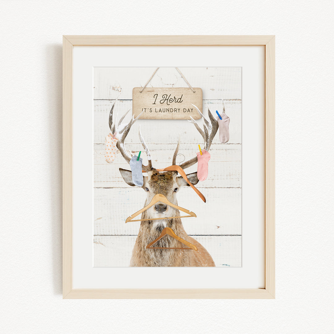 A framed image depicting a deer with antlers holding various laundry items, including socks and clothes, against a white background with a sign that reads "I herd its laundry day".