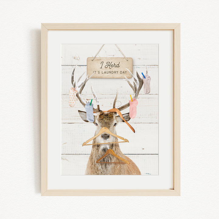 A framed image depicting a deer with antlers holding various laundry items, including socks and clothes, against a white background with a sign that reads "I herd its laundry day".
