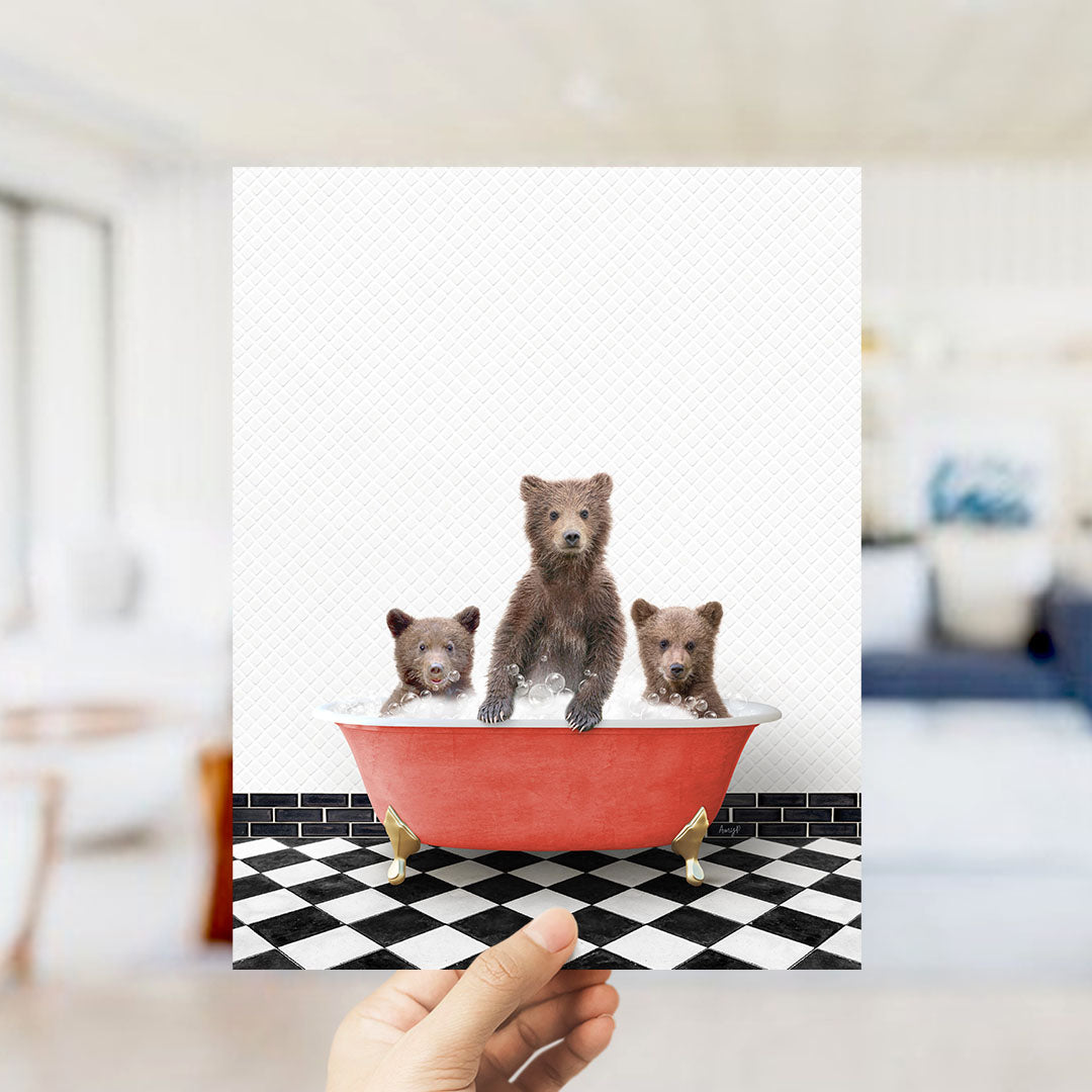 A hand holds up a framed image depicting three brown bears sitting in a red bathtub on a black and white checkered floor.