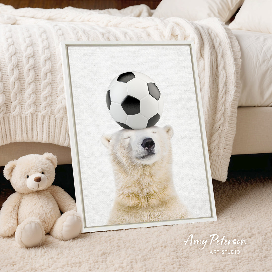 A framed image of a polar bear balancing a soccer ball on its head, with a teddy bear sitting next to it on a bed.