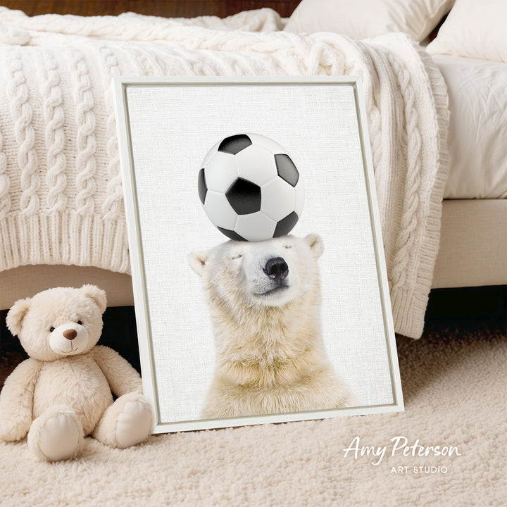 A framed image of a polar bear balancing a soccer ball on its head, with a teddy bear sitting next to it on a bed.