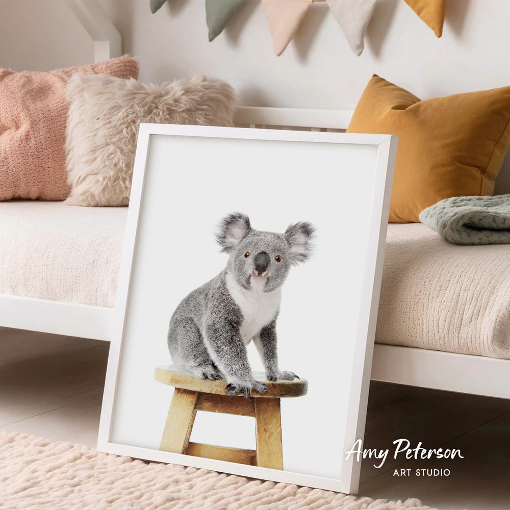 A framed photograph of a koala bear sitting on a wooden stool, displayed in a room with a white bed and colorful pillows.